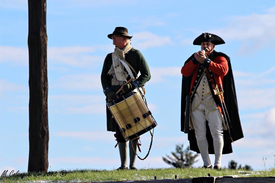 2 men stand on a grassy wall. One holds a drum. A second in a black cape, plays a bugle.
