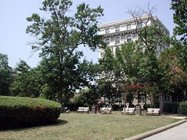 The east-west concrete walk located north of the memorial is lined with benches. The large canopy trees beyond partially obscure the historic building just north of them.