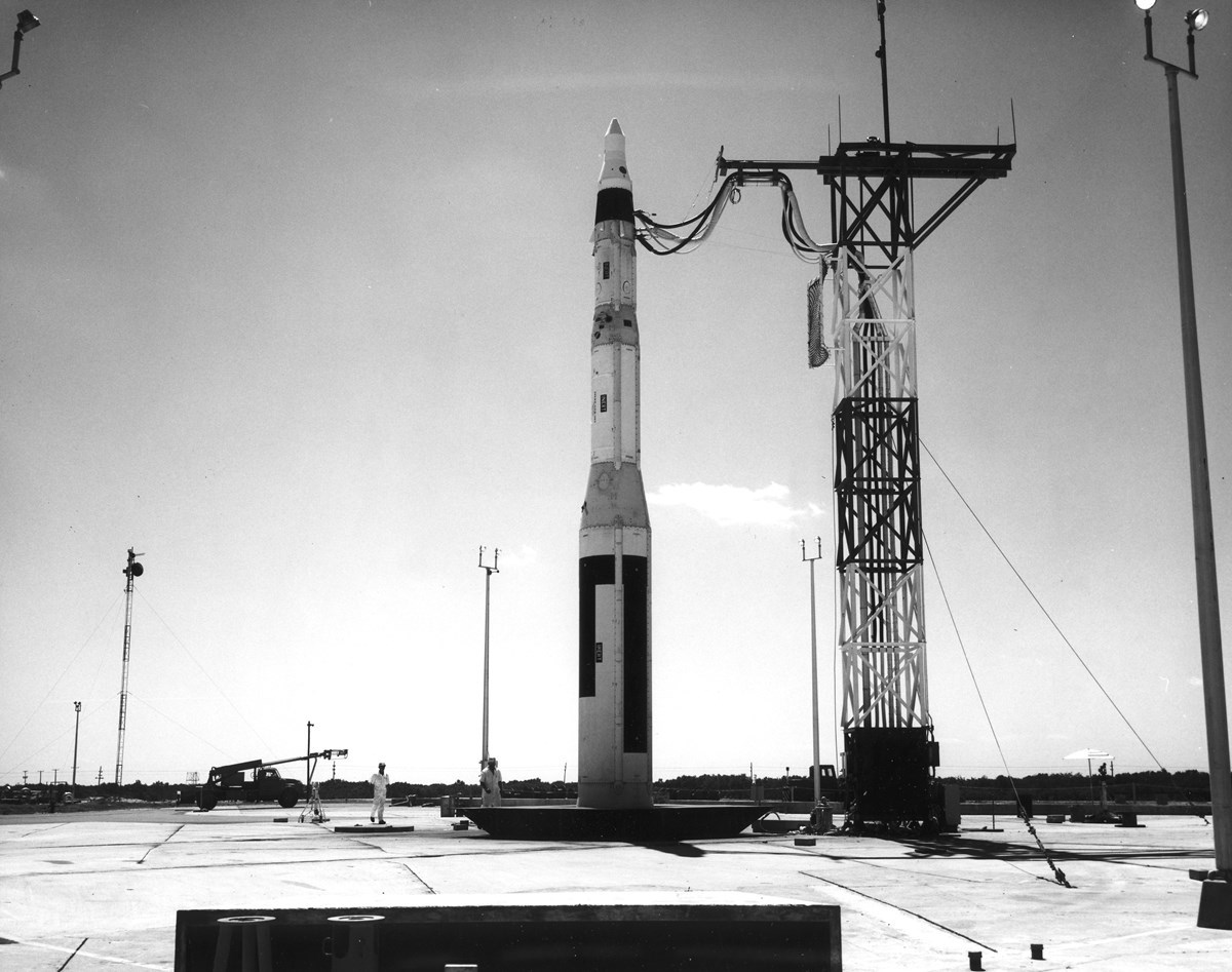 a small slender missile stands erect on a launch pad, connected to a tower
