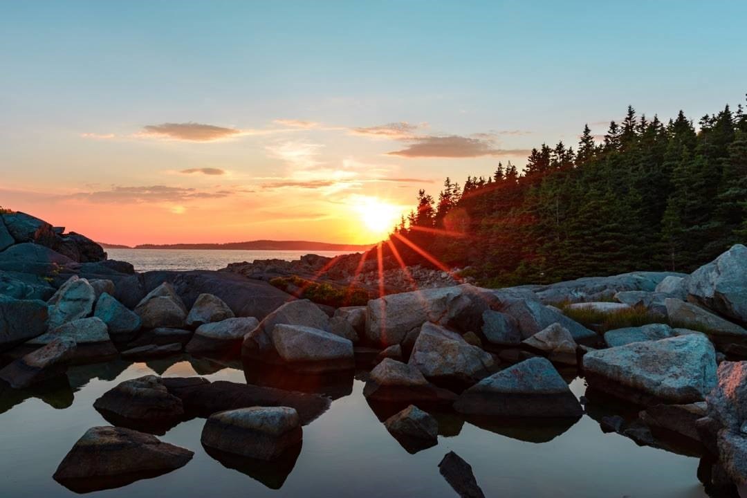 Sun setting over open water with rocks in the foreground and trees in the distance.