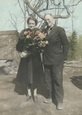 A man and a women standing next to one another outside. The women is holding a flower bouquet.