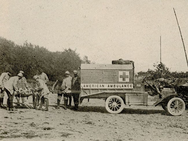 Group of men loading stretcher into back of truck marked with cross and "AMERICAN AMBULANCE"