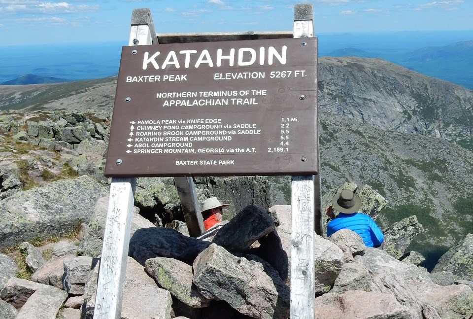 Backs of hikers behind a sign for Katahdin on a rocky mountain top