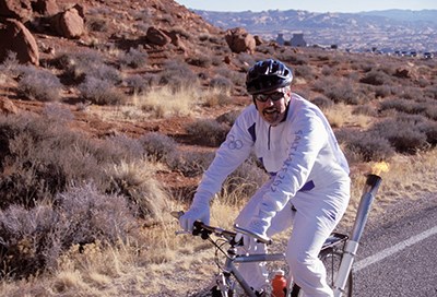 a cyclist in white Olympic uniform with a lit torch on the back of the bicycle