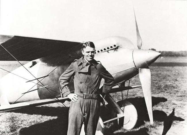 Black and white photo of man in flight suit standing in front of an aircraft.