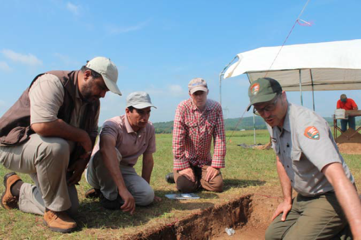 Andrew Weiland talking to foreign archeologists A ranger points to something in the ground while three other men watch from their knees