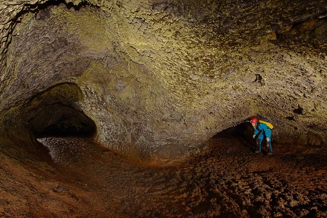 Geomicrobiologist in Hopkins Chocolate Cave at Lava Beds National Monument golden-brown interior cave wall with person wearing helmet and cave clothing crouched at its base