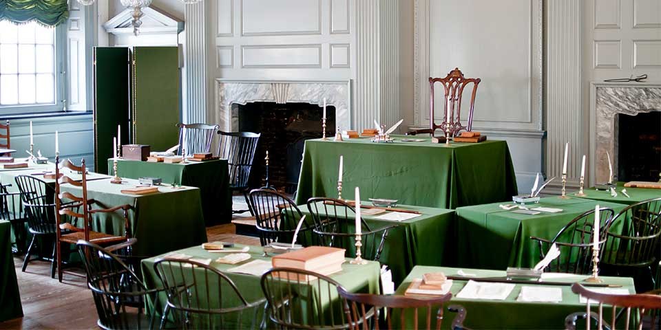Color photo of a room with rows of tables covered with green cloth facing a central table.
