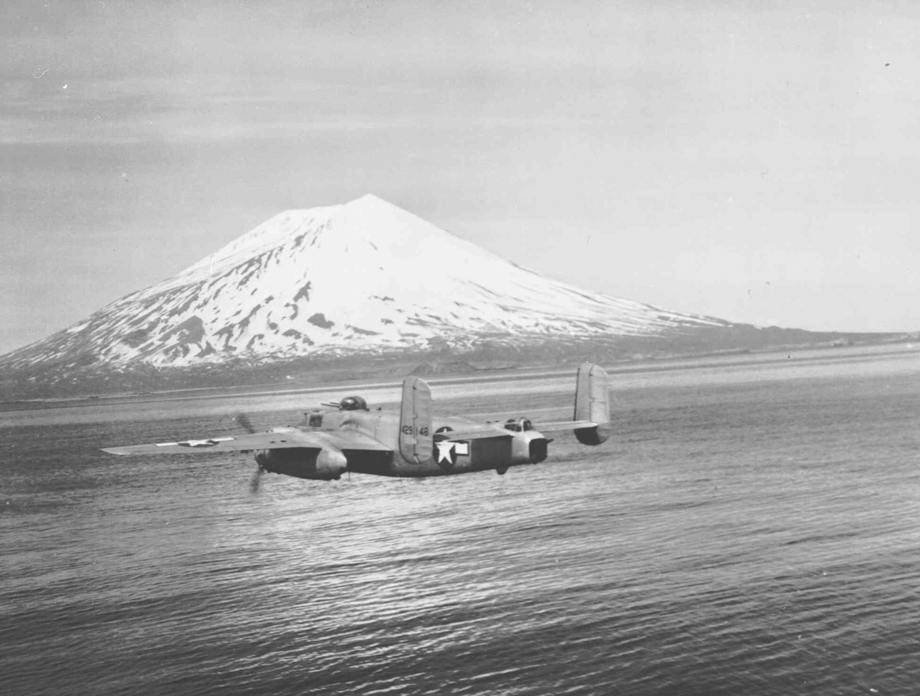 Plane flying over open water with snow-capped mountain in distance.