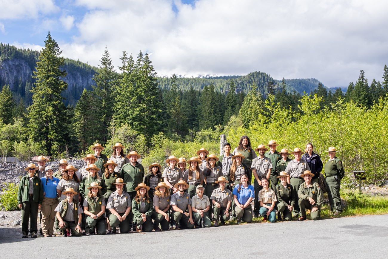 A large group of uniformed rangers pose in front of trees and clouds.