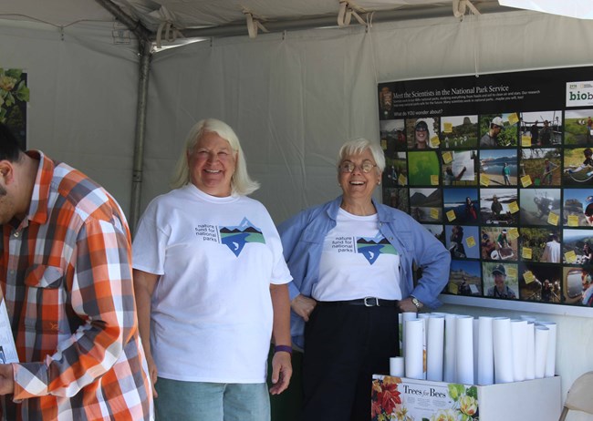 Chris Shaver and another Nature Fund for National Parks member smiling at from inside their booth, their friends group name across the top of it