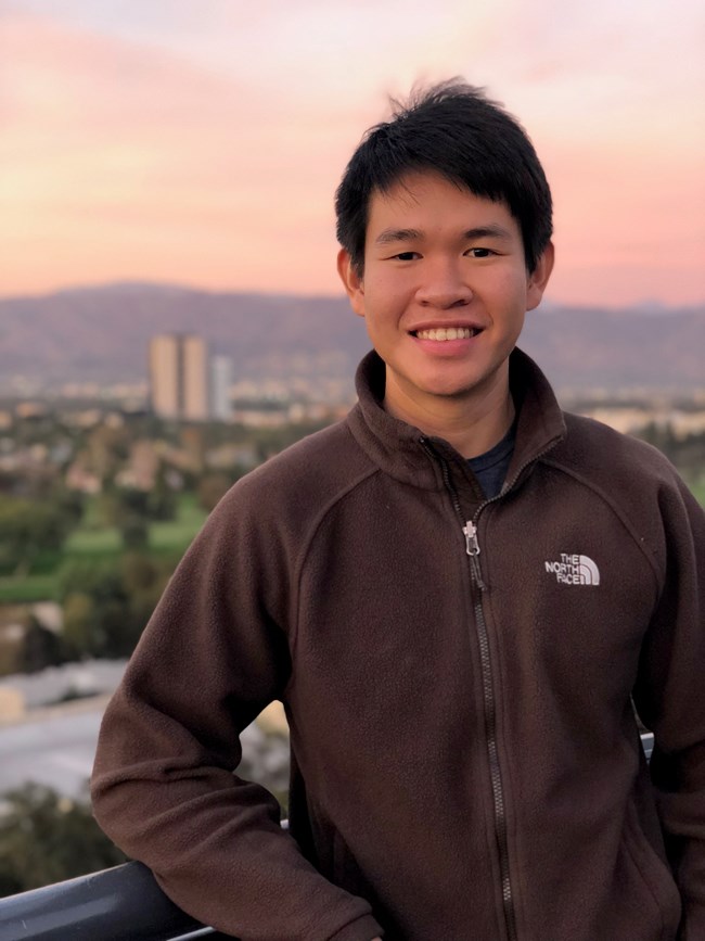 The author, Nuttapon (Nat) Pombubpa, smiling, in the background a cityscape with pink evening hues.