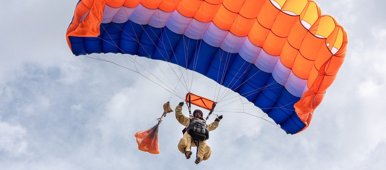 A smokejumper under a red, white, and blue rectangular canopy floats down from sunny skies.