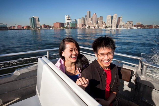 Two visitors sitting on a ferry out in open water with a cityscape in the background.