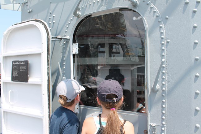 Children explore ships at the Boston Naval Yard. Two kids look through window of ship at Naval Yard.