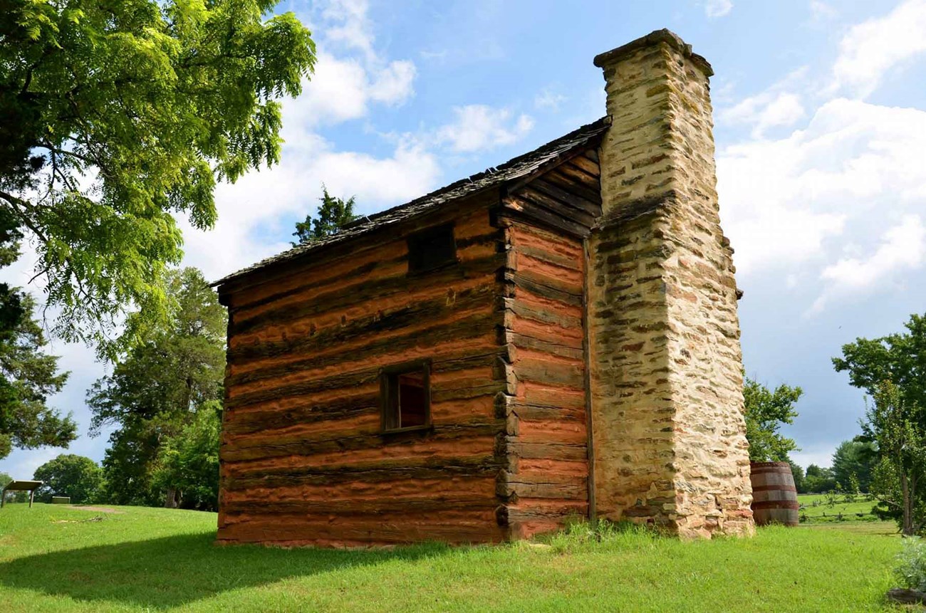 Log cabin with stone chimney surrounded by green grass and clear blue sky.