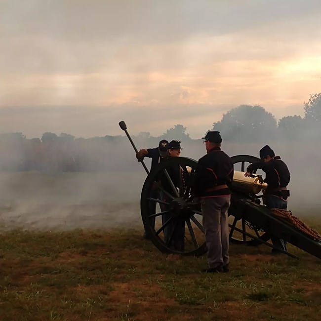 Color photo of 4 people standing around Civil War cannon in Civil War uniform. There is a slight fog around them as the sun begins to set