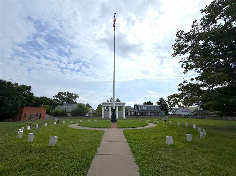 A modern photo of a cemetery with a flagpole surrounded by gravestones with buildings, trees, green grass, blue sky with clouds