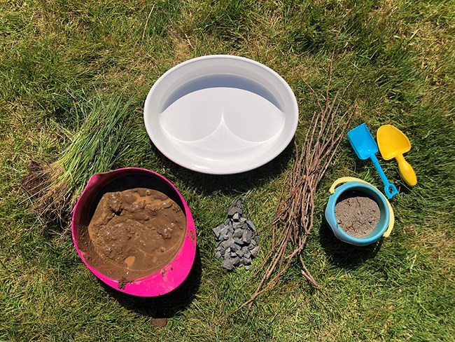 Photo of supplies laid out on grass: a baking dish, bowl of mud, grass, rocks, sticks, and plastic shovels.