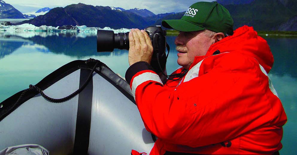 A glaciologist takes photos of a glacier while wearing a large, bright red winter coat.