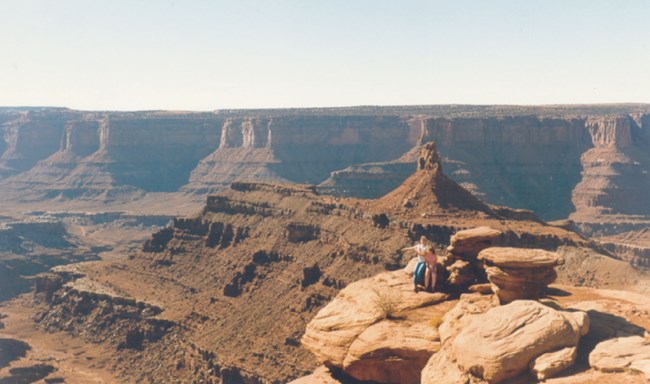 2 people on a canyon rim above deeply eroded river canyon.