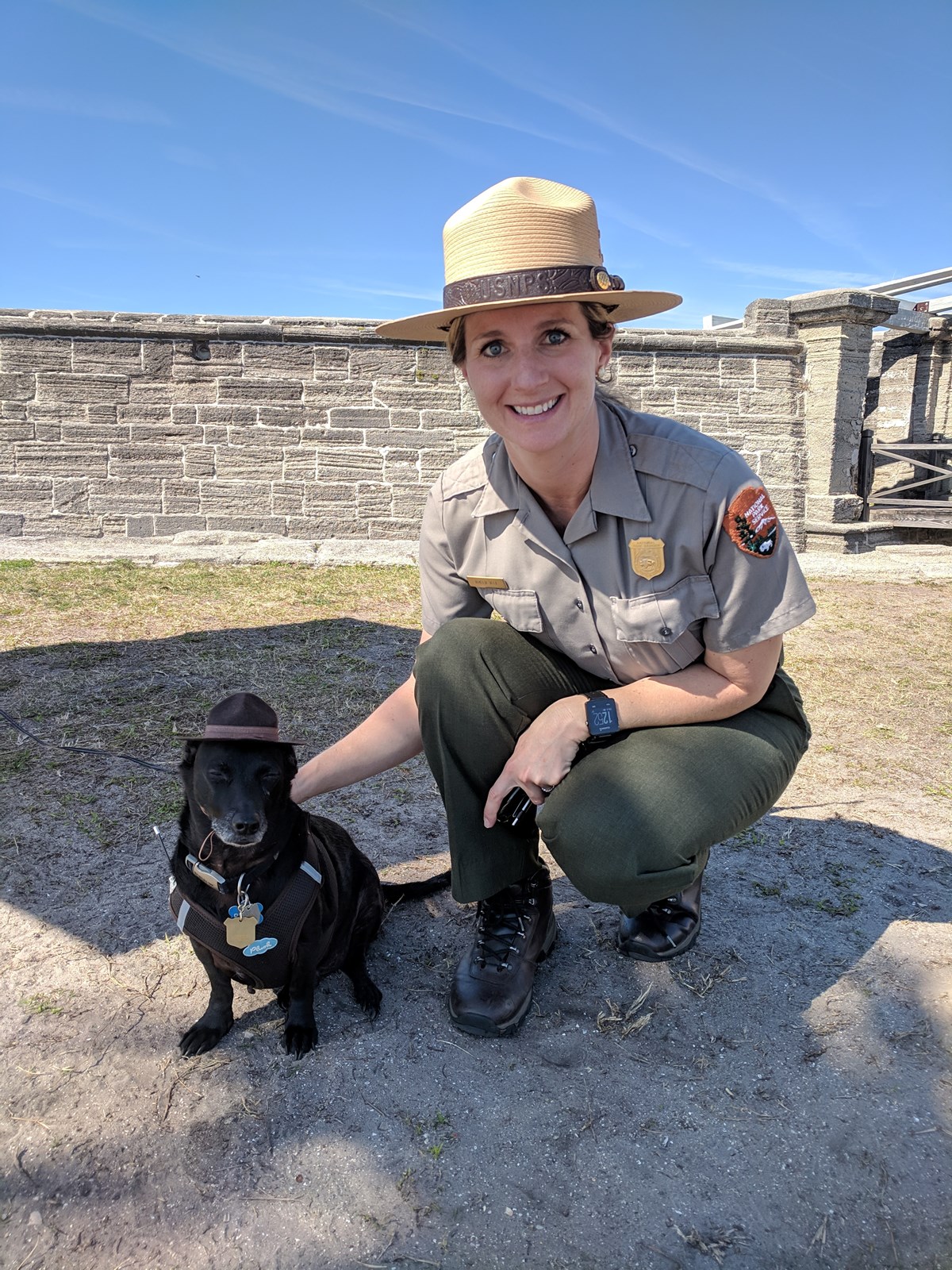 Image of a dog with ranger hat and badge with a Park Ranger outside of the fort called, Castillo de San Marcos.