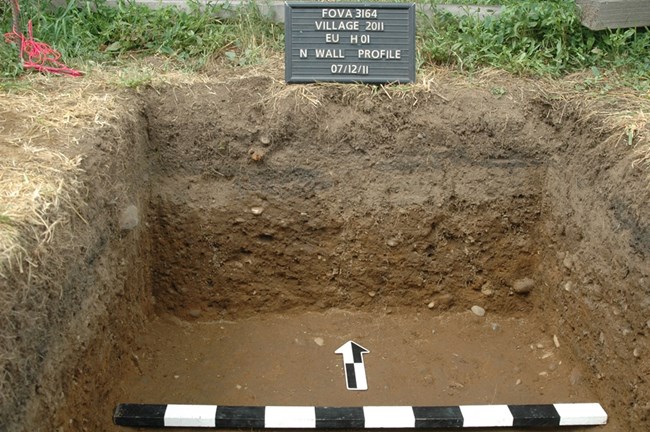 Photo of excavation unit. A dark grey stripe around the unit's walls shows where the road once was.