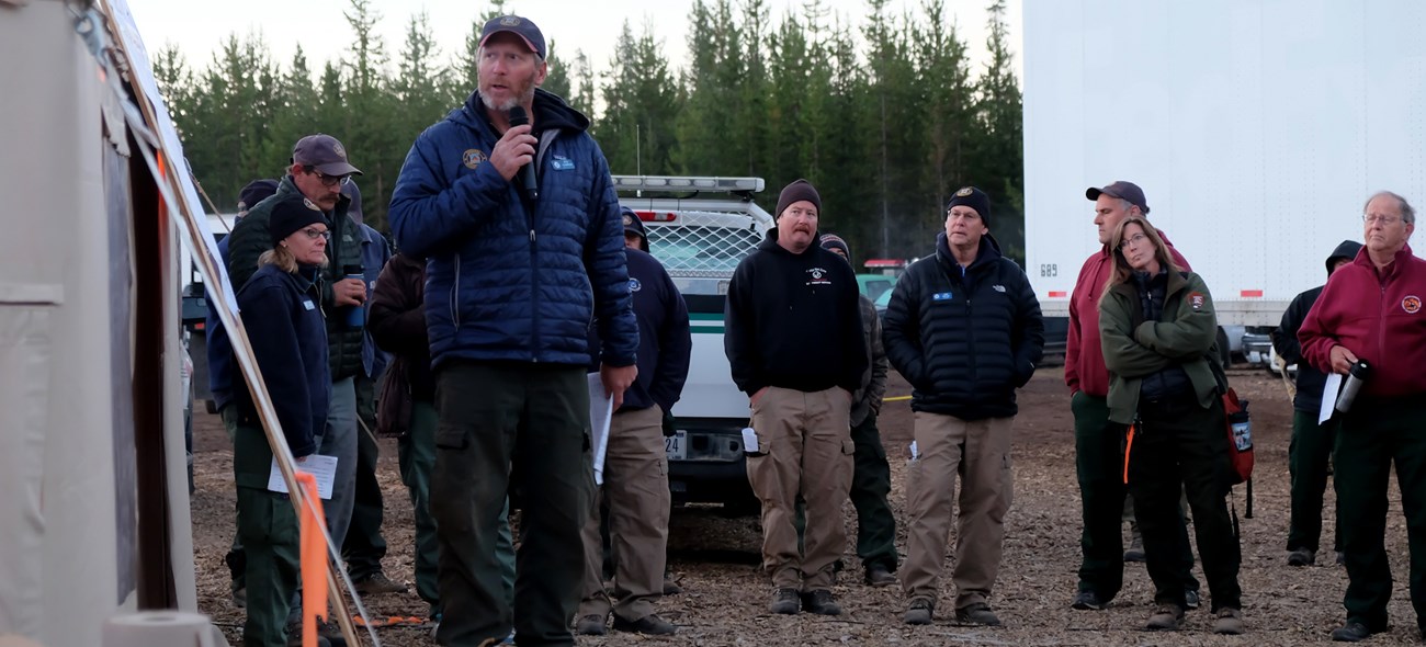 Briefing on the Bybee Creek Fire at Crater Lake National Park. A man stands near a board in front of a group of people speaking into a microphone.