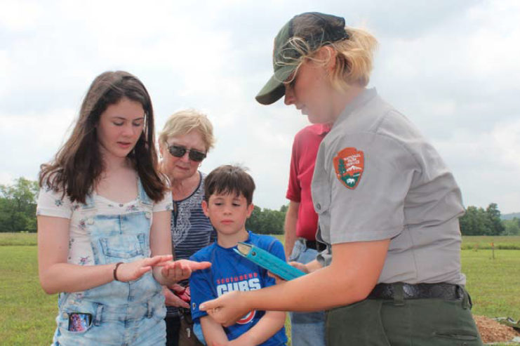 Cailey Mullins teaching kids about archy A ranger holding a blue book and interacting with kids and adults.