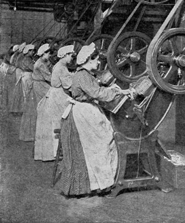 Row of women in white caps sitting at a row of machines in a factory