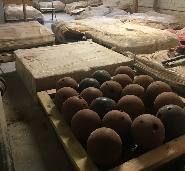 Crates of cannonballs and other historic materials fill a brick walled room.