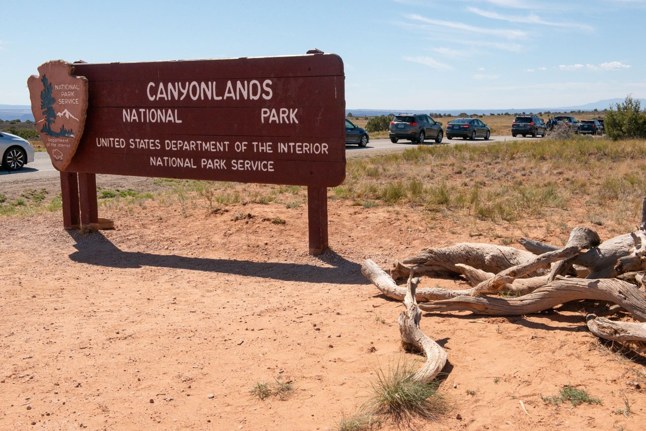 Several cars line the road to enter Canyonlands Island in the Sky District. The Entrance sign is visible in the foreground.