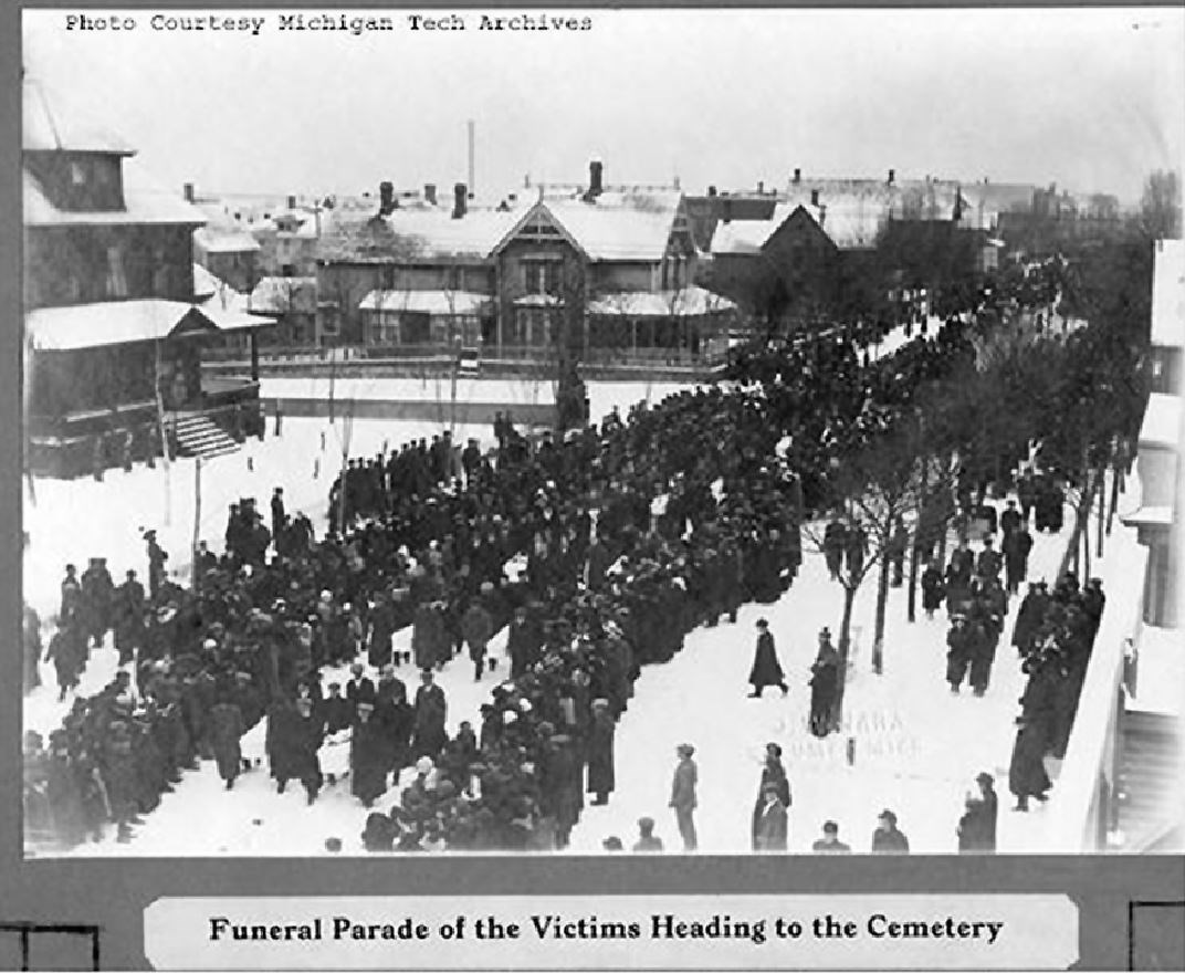 Crowds of people walking through streets of a town with words "Funeral Parade of Victims Heading to Cemetery" printed on bottom of photograph.