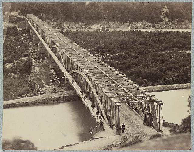 Sepia tone photo of Chain Bridge crossing the Potomac river and C&O Canal. Taken from up high showing men walking, fishing, and posing for the photo. The land under the bridge is dense with vegetation and rocky shores