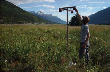 a person reaches up to adjust a gas measuring device in a grassy field, surrounded by mountains