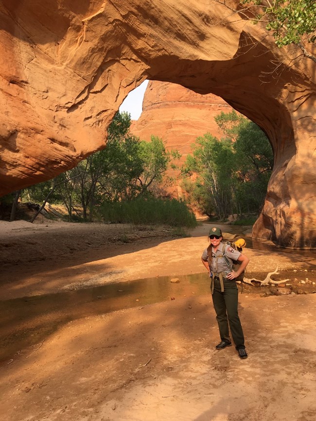 lt= A park ranger stands under a tall red rock arch.
