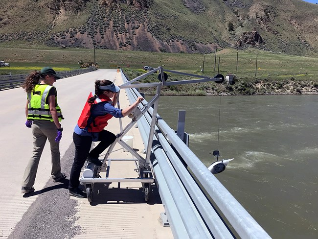 A suspension sampler being lowered with a reel and bridge board from a bridge over the Yellowstone River Two researchers on a bridge over a river lowering sampler from a bridge board