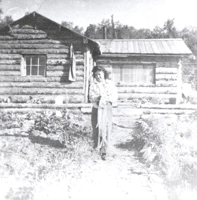 Mary Alsworth in front of Kennedy Cabin, c. 1942 Historic black and white image of a woman standing in front of log cabin.