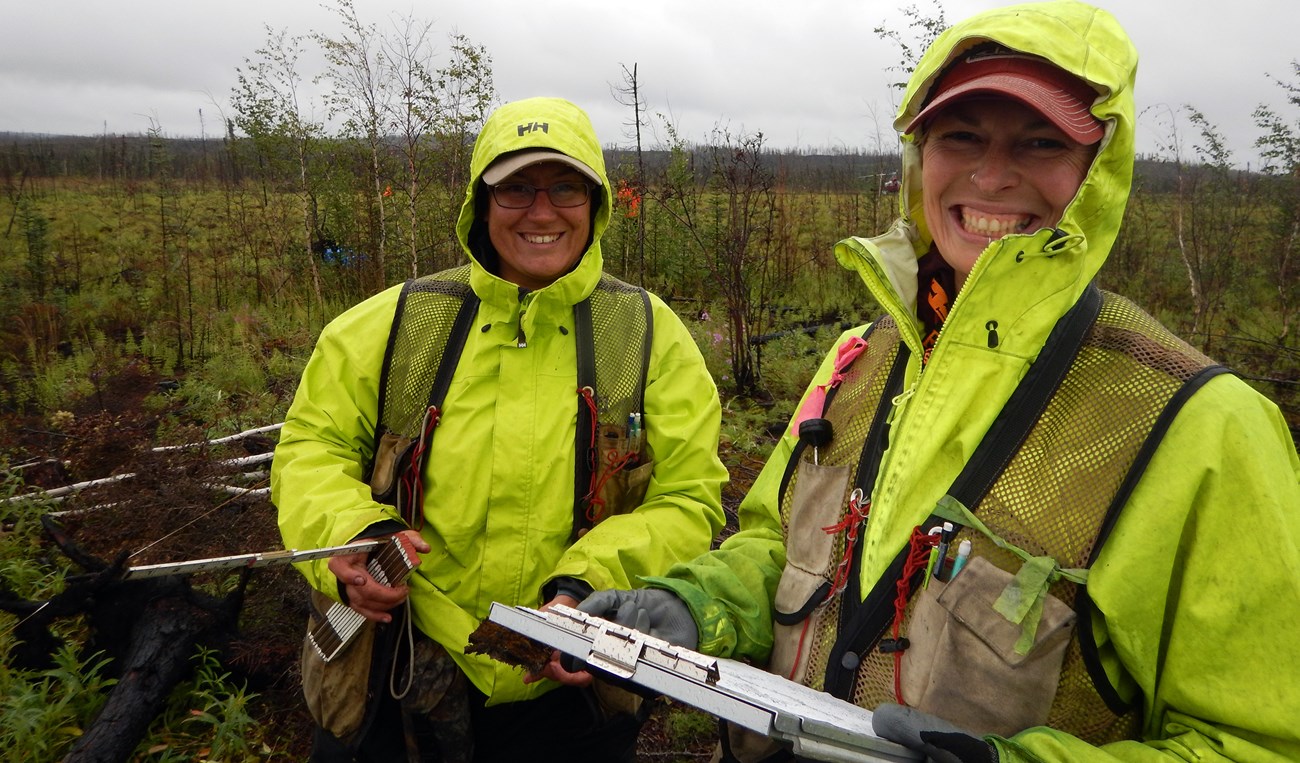Two people with neon green rain jackets hold paperwork and a measuring stick.