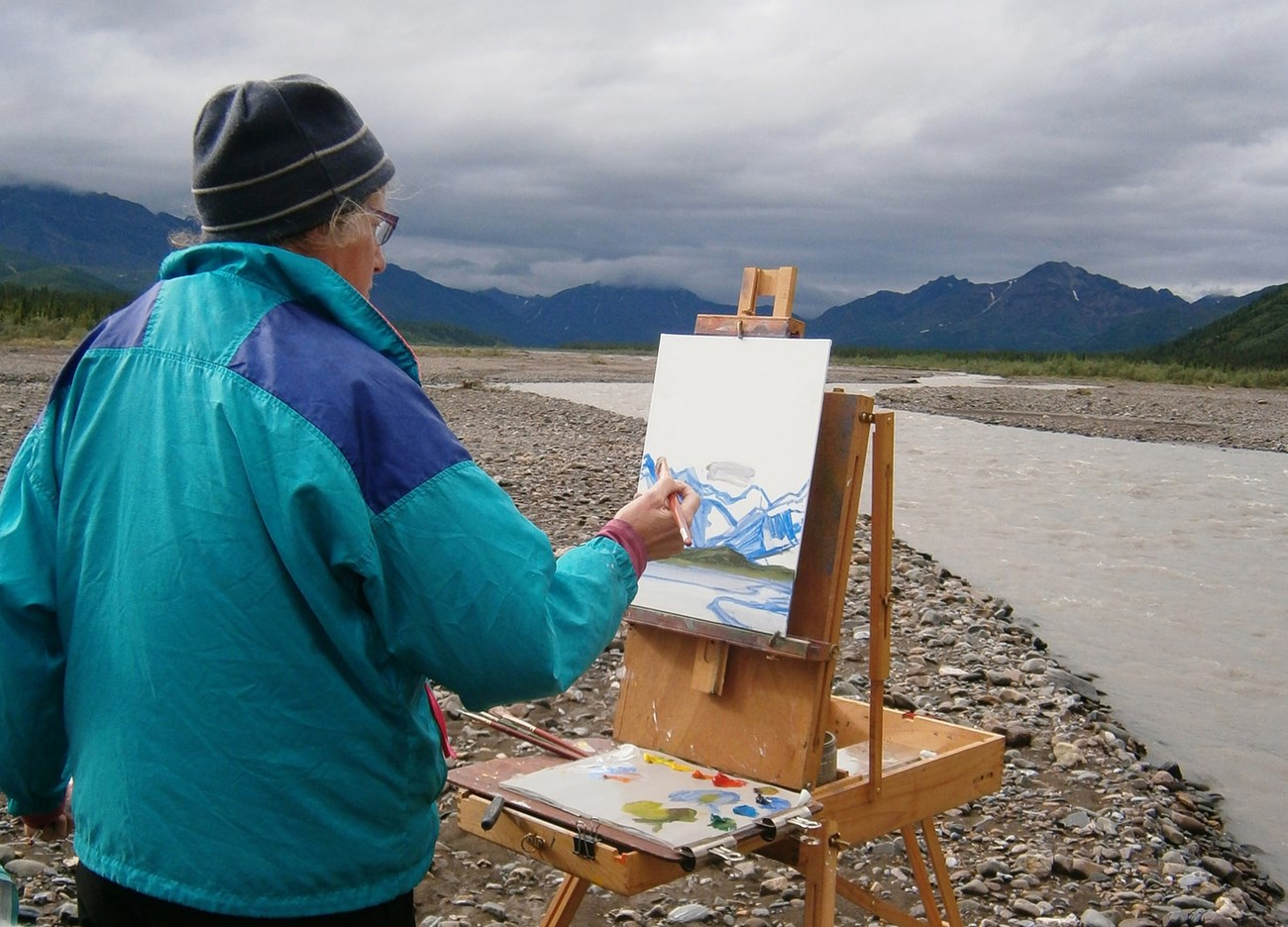 Man painting a canvas on an easel along a shoreline.