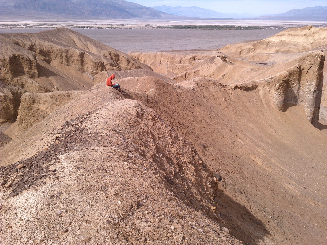 A researcher sits on a narrow, steep ridge of rocky, tan-colored terrain, writing down observations.