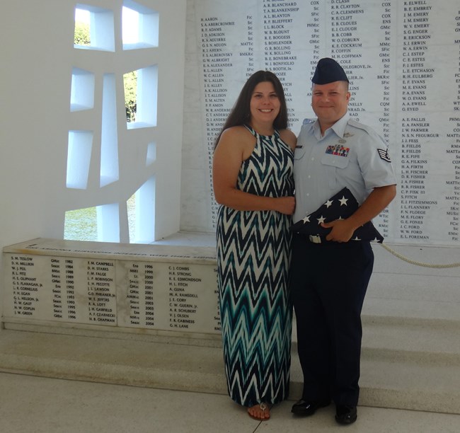 Woman in dress locks arms with a man in Air Force uniform holding a folded flag. They stand in front of the USS Arizona Memorial wall.