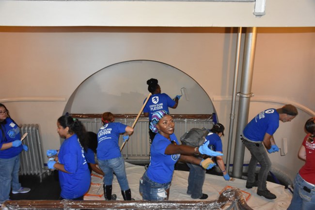 Eight people in blue shirts each in various stages of painting walls.