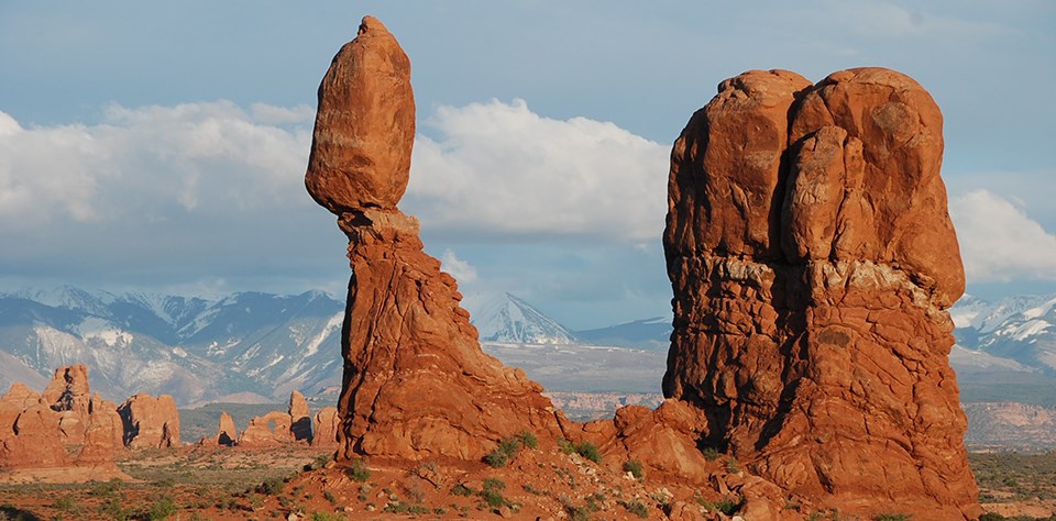 a tall balanced rock with a distant arch and snow-capped mountains in the background