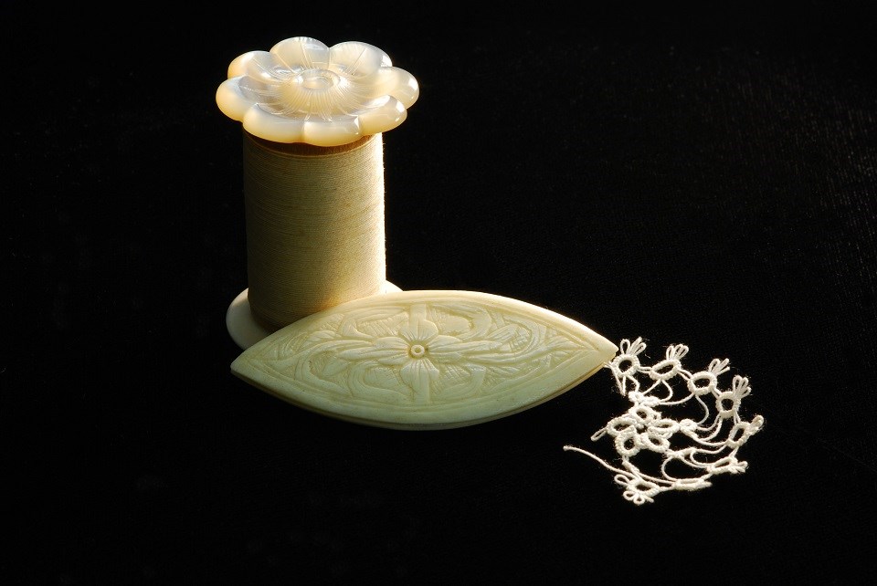 Photo of spool of white thread with decorative top. Next to the spool is a flat almond-shaped tool with some tatting in white thread attached to it.