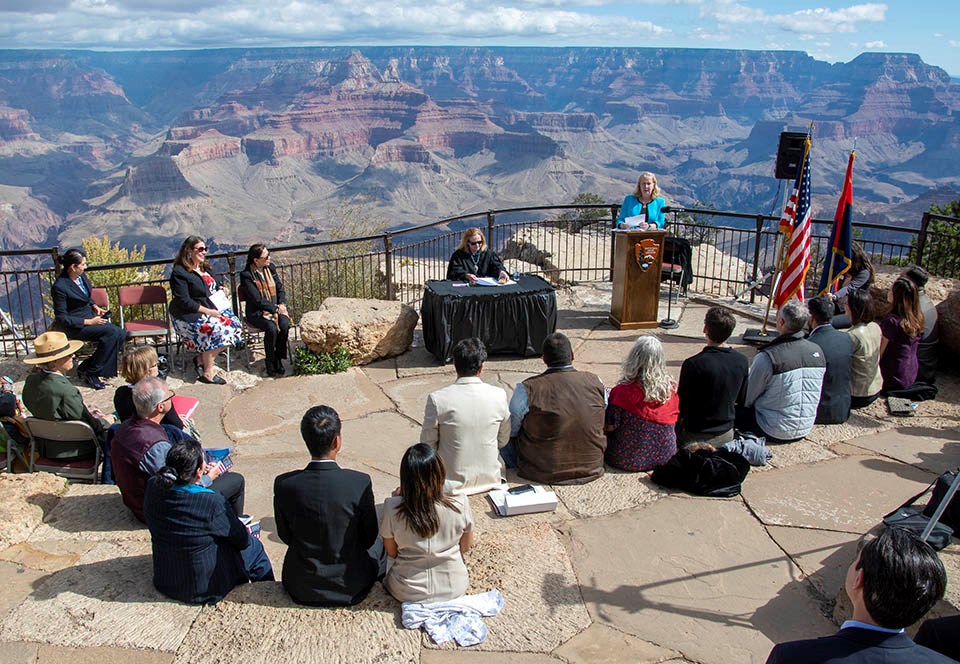 Naturalization Ceremony at Mather Amphitheater on the South Rim of Grand Canyon National Park. 15 people sit in a semicircle in a stone amphitheater. A judge in black robes is sitting at a table with a black tablecloth. A Woman dressed in blue is behind a podium and addressing the group. In the distance, a colorful landscape of peaks and cliffs.