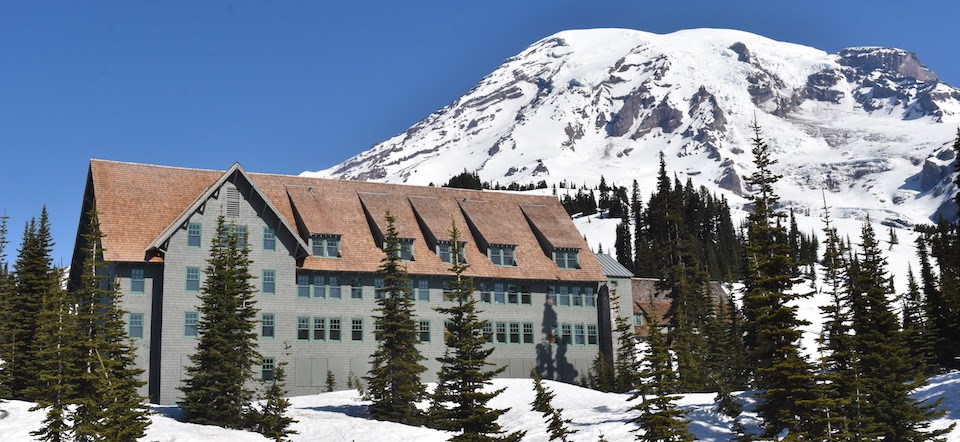 A four story wood building with an angled roof in front of a glaciated mountain.