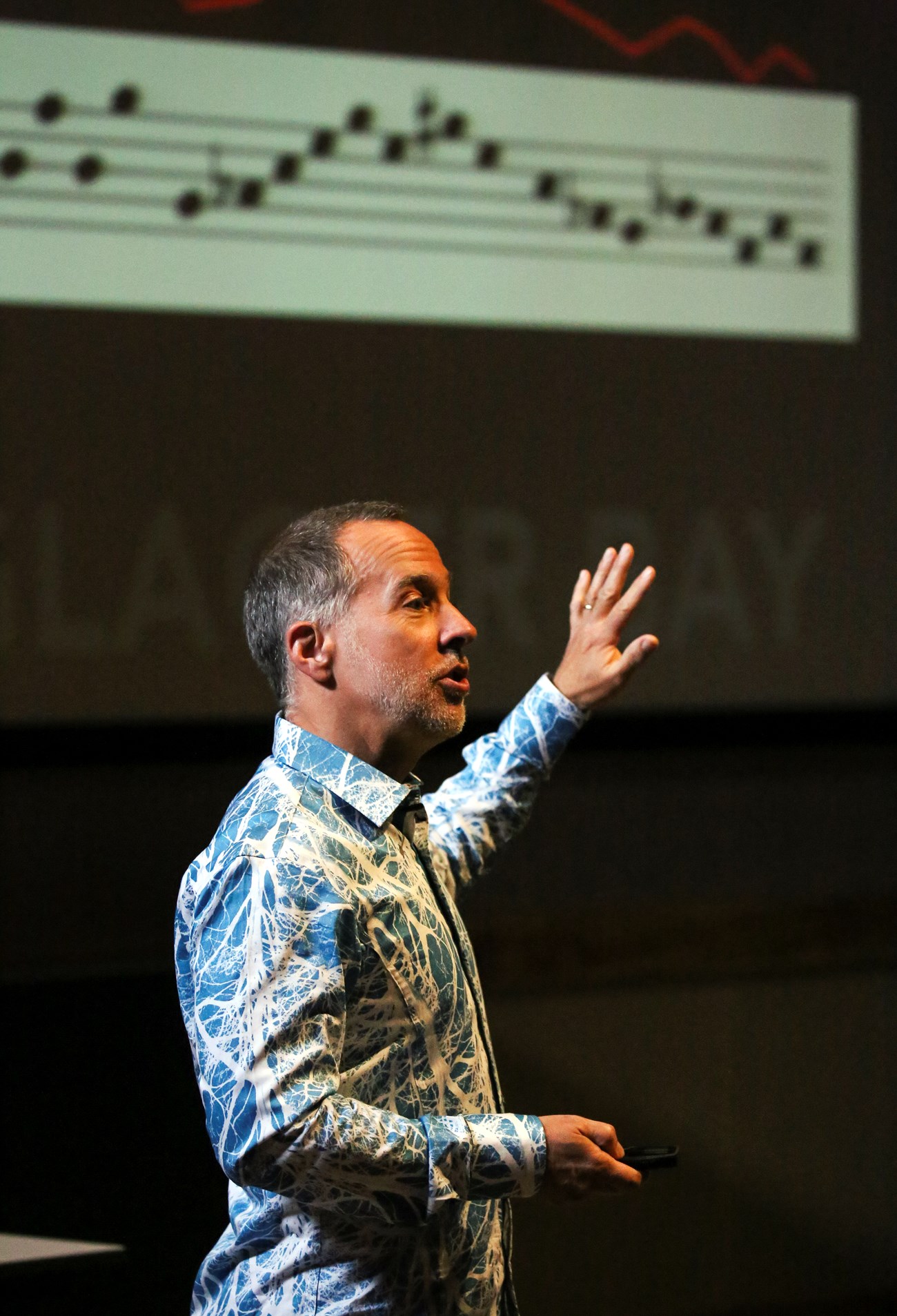 man in an auditorium gesturing in front of a screen showing music notes
