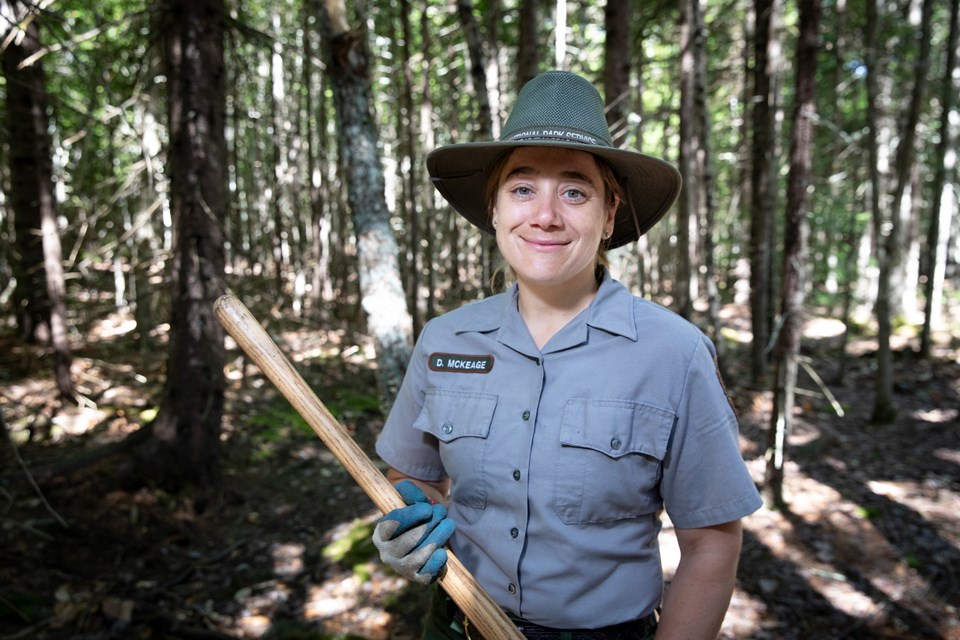 Woman wearing a broad brim work hat and gloves holds the wood handle of a trail building tool