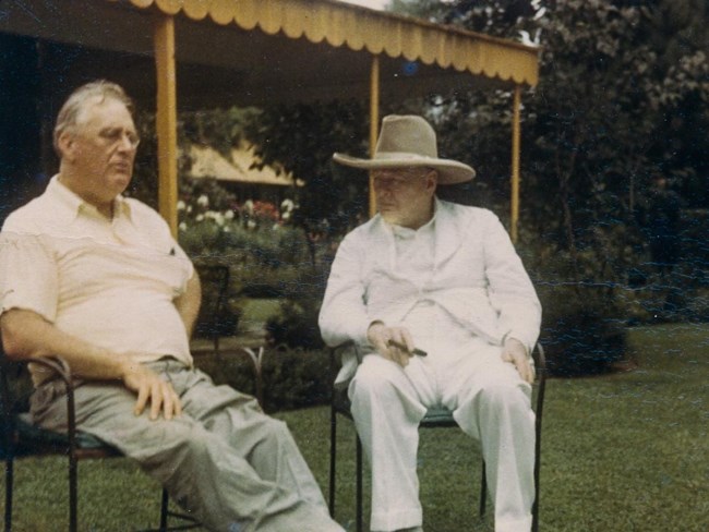 lt= Two men seated on a lawn in conversation.